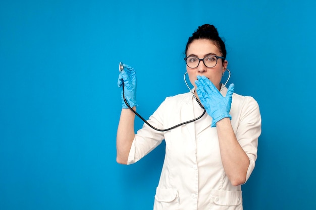 Médica chocada de uniforme usa estetoscópio e escuta em fundo azul