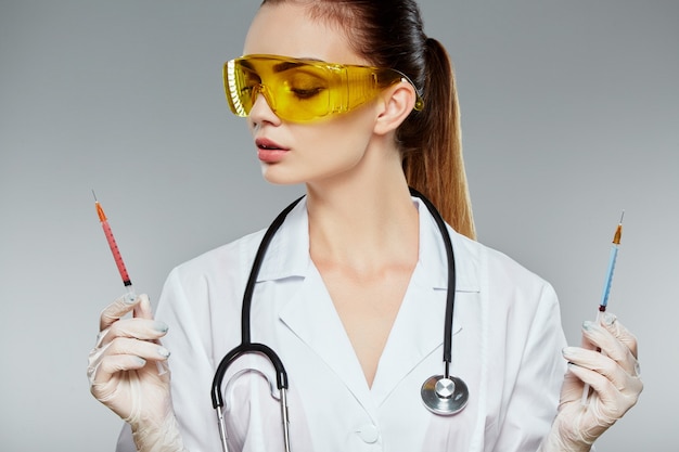 Médica com cabelo castanho e nude maquiagem vestindo uniforme médico branco, óculos, estetoscópios e luvas brancas no fundo cinza do estúdio e segurando seringas.