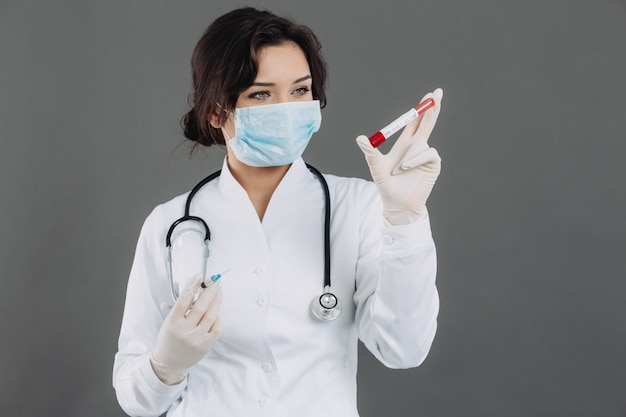 Médica com estetoscópio na máscara segurando a injeção, antídoto contra o coronavírus em fundo cinza.