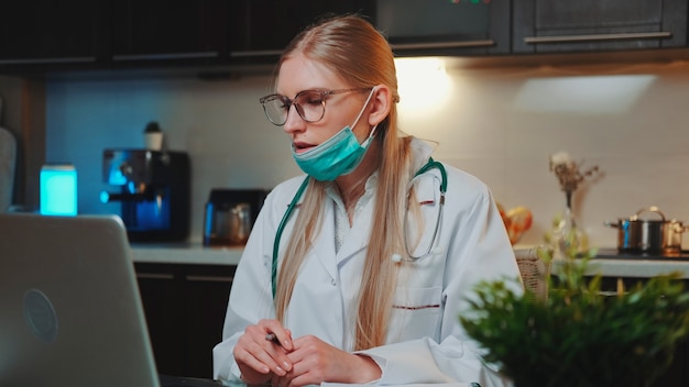 Médica com máscara médica em vestido de médico, fazendo videochamada para paciente de casa em que ela está sentada ...