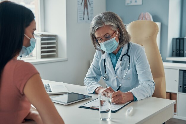 Médica confiante, com jaleco branco, prescrevendo uma receita para uma jovem sentada em seu escritório