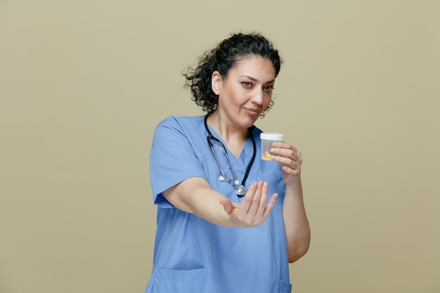 médica confiante de meia-idade vestindo uniforme e estetoscópio no pescoço segurando o recipiente de medição com comprimidos nele olhando para o lado mostrando venha aqui gesto isolado no fundo verde-oliva