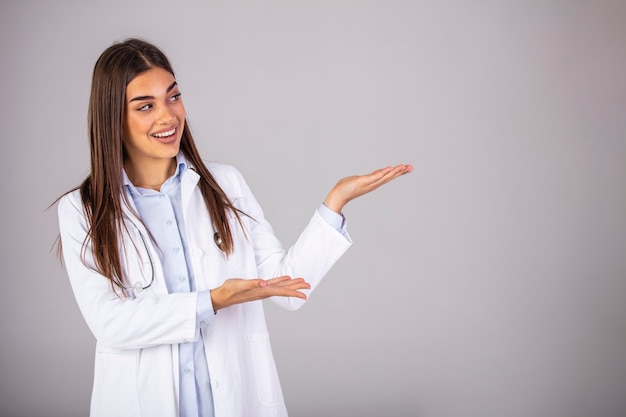 Médica confiante em uniforme branco, olhando para longe e apontando em pé contra um fundo cinza. olhe para lá! aqui está o que seu médico recomenda.