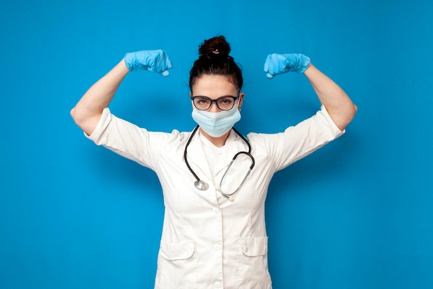 Médica de bata médica e máscara médica mostra gesto de força em fundo azul