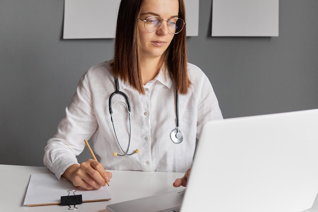 Médica de cabelos escuros trabalhando em uma mesa de escritório usando óculos e bata de laboratório branca olhando para a tela do laptop escrevendo preenchendo o histórico médico do paciente