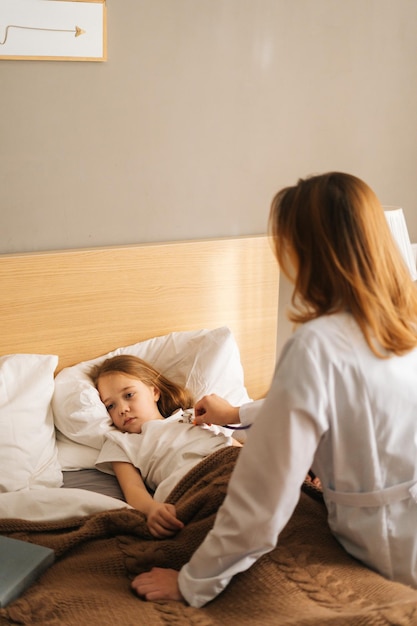 Médica de jaleco branco ouvindo batimentos cardíacos com estetoscópio para verificar menina doente deitada na cama no hospital