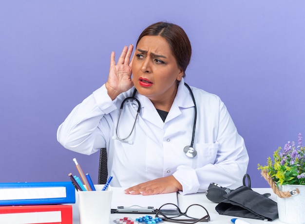 Médica de meia-idade com jaleco branco com estetoscópio olhando para o lado com a mão na orelha tentando ouvir, sentada à mesa sobre a parede azul