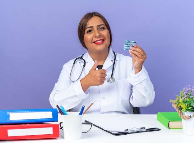 Médica de meia-idade com jaleco branco com estetoscópio segurando comprimidos, olhando para a frente, feliz e positiva, sorrindo, mostrando os polegares para cima, sentado à mesa sobre a parede azul