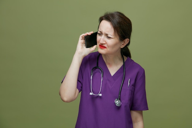 Médica de meia-idade irritada vestindo uniforme e estetoscópio no pescoço, falando no telefone, olhando para o lado isolado no fundo verde-oliva