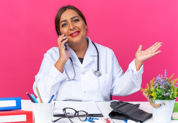 Médica de meia-idade vestindo jaleco branco com estetoscópio e sorrindo confiante enquanto fala no celular, sentada à mesa com pastas de escritório sobre fundo rosa