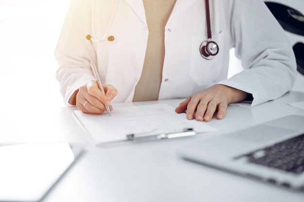 Médica desconocida sentada y escribiendo notas en el escritorio de una clínica o oficina del hospital, de cerca.