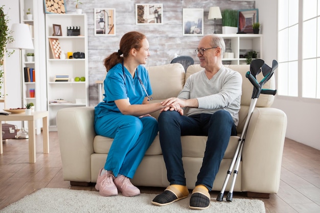 Médica e homem sênior sorrindo um para o outro enquanto está sentado no sofá no lar de idosos.