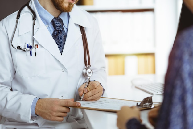 Médica e paciente do sexo masculino discutindo o exame de saúde atual enquanto está sentado na clínica ensolarada, close-up.