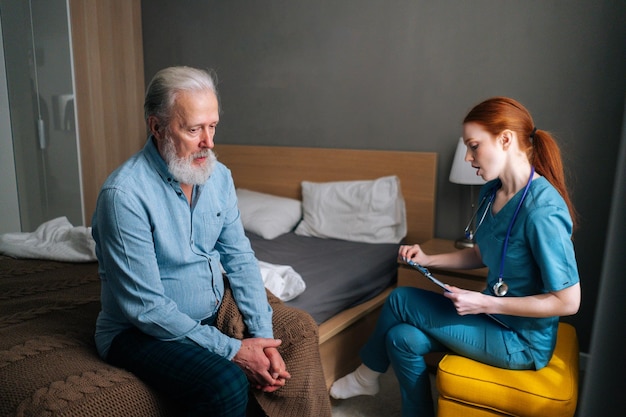 Médica falando com paciente do sexo masculino adulto sênior sentado na cama em casa e verificando os sintomas de escrita de saúde no histórico médico na área de transferência