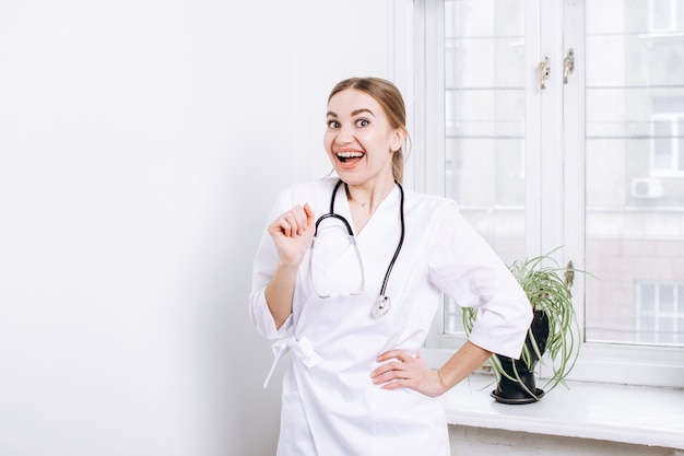 Médica feliz com um jaleco branco e um estetoscópio perto da janela