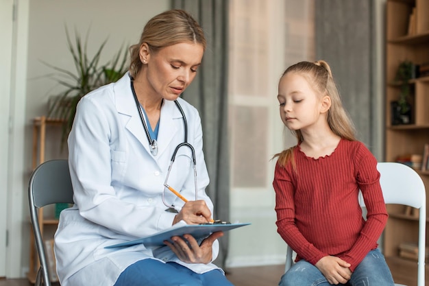 Médica focada anotando os sintomas da menina na área de transferência enquanto criança olhando o que está escrevendo no gráfico