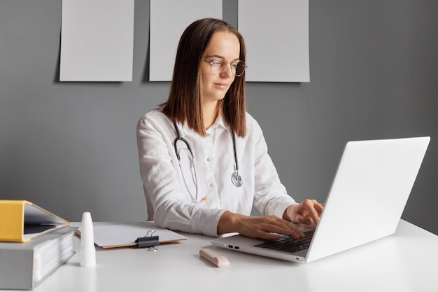 Médica focada e concentrada vestindo uma bata de laboratório sentada à mesa e trabalhando em um laptop com base de dados eletrônica digitando no teclado olhando para a tela