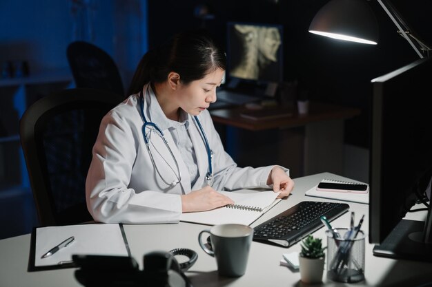 médica japonesa asiática sentada à mesa e escrevendo no relatório do documento no escritório do hospital à noite. Conceito de serviço de equipe de saúde médica. mulher trabalhadora enfermeira lendo caderno