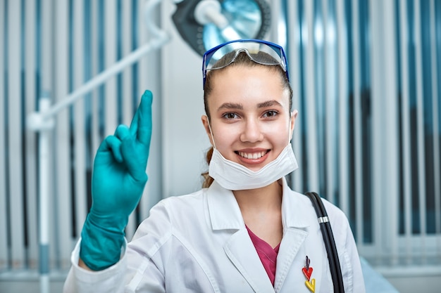 Médica jovem bonita com uniforme branco e máscara no rosto com dedos cruzados no fundo da clínica