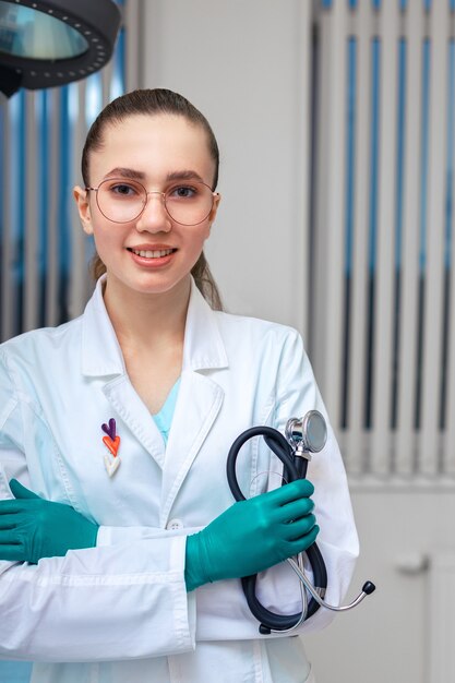 Médica jovem com um casaco e um estetoscópio nas mãos, posando para a câmera na superfície da enfermaria do hospital