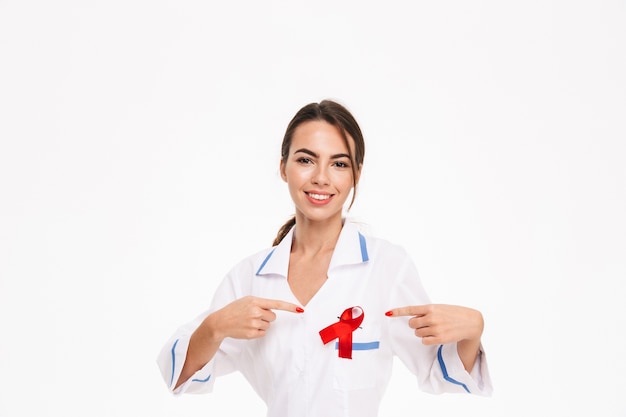 Médica jovem sorridente usando uniforme com uma fita vermelha isolada na parede branca