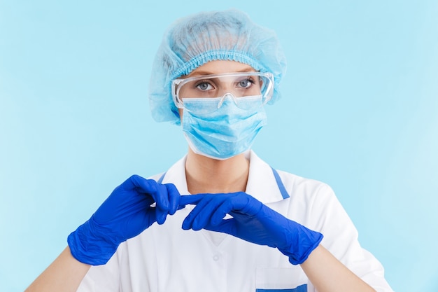 Médica linda mulher loira confiante usando uniforme e máscara em pé isolado sobre a parede azul, preparando-se para um procedimento