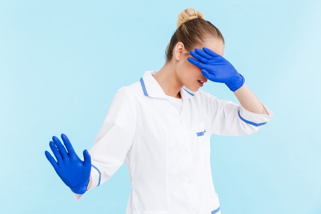 Médica loira linda vestindo uniforme em pé, isolado na parede azul, cobrindo o rosto com a mão