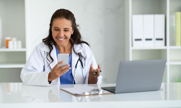 Foto médica madura e alegre sentada em seu armário usando telefone