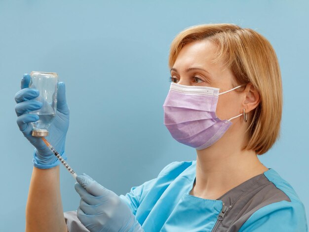 Médica con mascarilla y guantes sosteniendo una botella de vidrio y llenando una jeringa para inyección