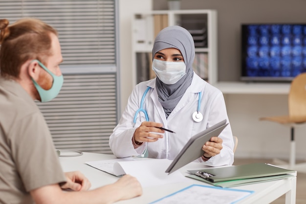 Médica muçulmana apontando para um tablet digital e discutindo problemas de saúde com seu paciente no hospital