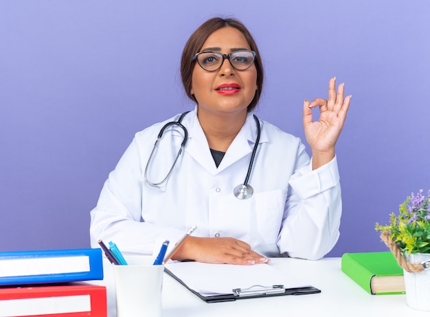 Médica mulher de jaleco branco com estetoscópio e óculos, olhando para a frente, sorrindo confiante fazendo um sinal de ok sentado à mesa sobre a parede azul