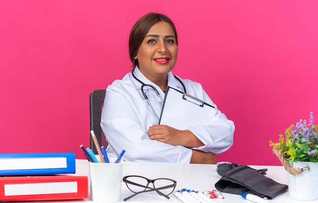 Médica mulher de jaleco branco com estetoscópio segurando a prancheta e sorrindo confiante, sentada à mesa com pastas de escritório sobre fundo rosa