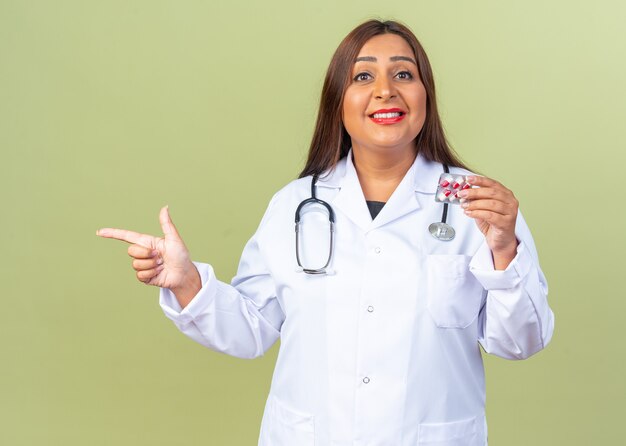 Foto médica mulher de jaleco branco com estetoscópio segurando uma bolha com comprimidos apontando com o dedo indicador para o lado sorrindo confiante em pé no verde