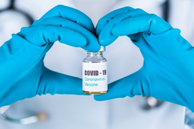 Médica ou enfermeira de uniforme e luvas usando máscara protetora em laboratório segurando uma forma de frasco de vacina de medicamento para o coração com rótulo de vacina de Coronovírus COVID-19