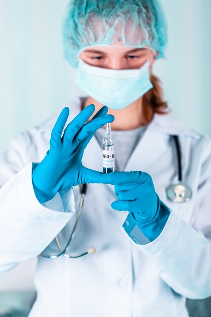 Médica ou enfermeira de uniforme e luvas usando máscara protetora no laboratório, segurando o frasco de vacina de medicamento com rótulo de vacina de Coronovírus COVID-19