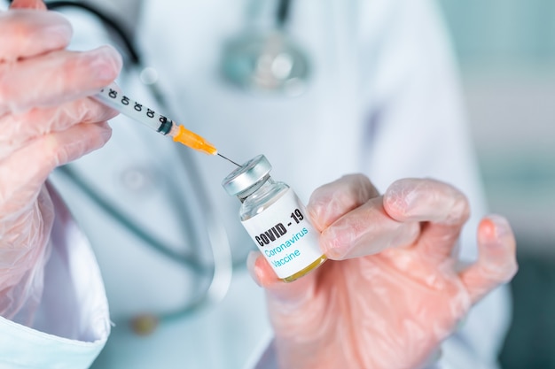 Médica ou enfermeira de uniforme e luvas usando máscara protetora no laboratório, segurando um frasco de vacina de medicamento com rótulo de vacina de coronovírus covid-19