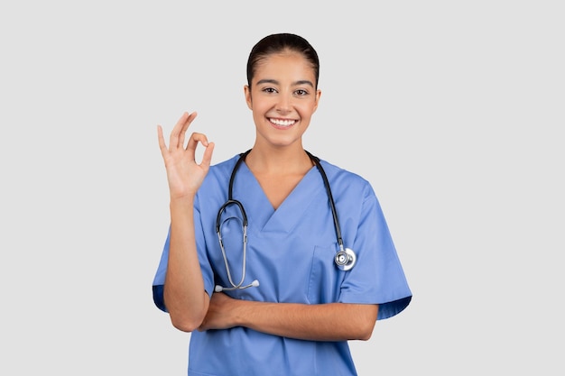Médica ou enfermeira milenar caucasiana positiva em uniforme azul mostra sinal ok com a mão