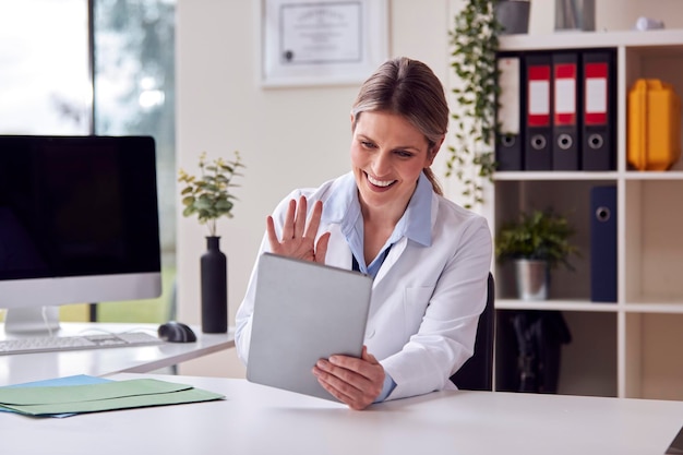 Médica ou GP vestindo jaleco branco sentado na mesa no escritório tendo chamada de vídeo no tablet digital