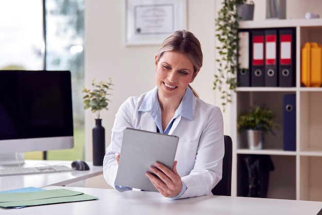 Médica ou GP vestindo jaleco branco sentado na mesa no escritório usando tablet digital