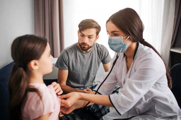 Médica ouvir respiração da menina pequena. O pai dela se senta e olha para a filha.