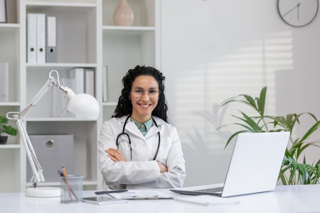 Médica profesional hispana con los brazos cruzados sentada en su escritorio con una computadora portátil y