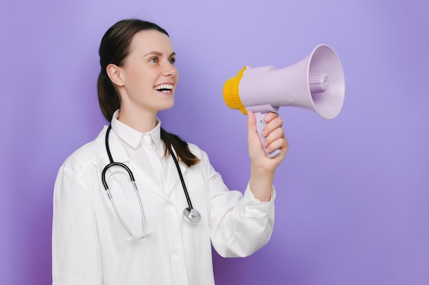 Foto médica, profissional médica se comunica gritando alto segurando o megafone, expressando sucesso e conceito positivo, ideia para marketing ou vendas, posando isolado sobre o fundo roxo do estúdio