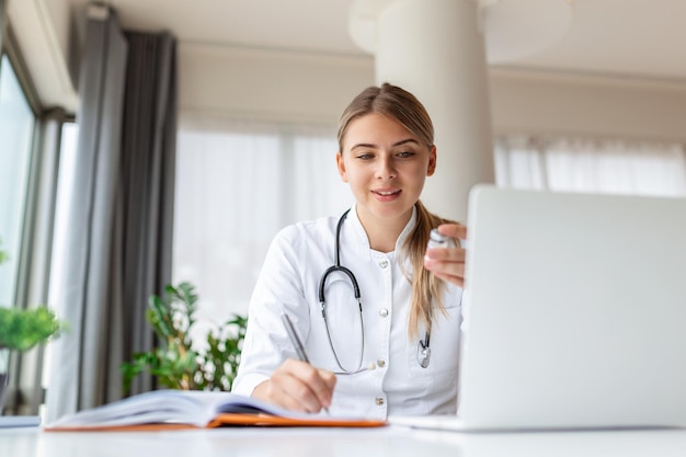Médica profissional sorridente vestindo uniforme tomando notas no diário médico preenchendo documentos história de doença do paciente olhando para tela do laptop estudante assistindo webinar