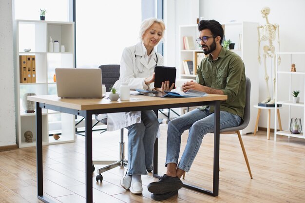 Foto médica sênior e paciente masculino sentados na mesa de um centro médico moderno