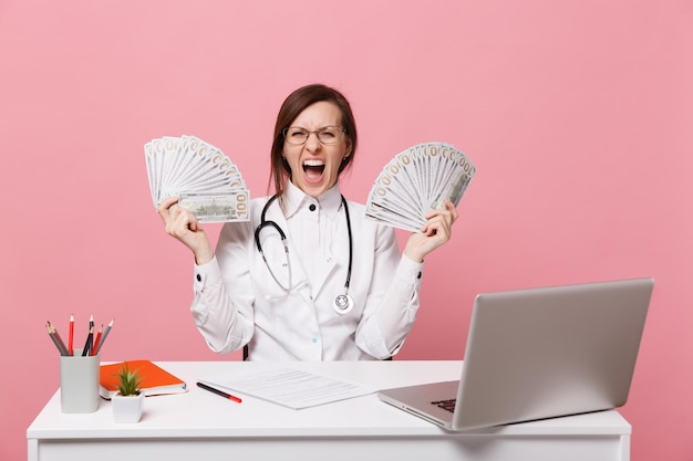Médica senta-se na mesa de trabalho no computador com dinheiro de documento médico no hospital isolado no fundo da parede rosa pastel. Mulher de estetoscópio de óculos de vestido médico. Conceito de medicina de saúde