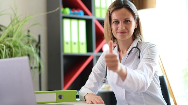 Médica sentada em sua mesa na clínica médica e mostra polegar para cima trabalhador médico olhando