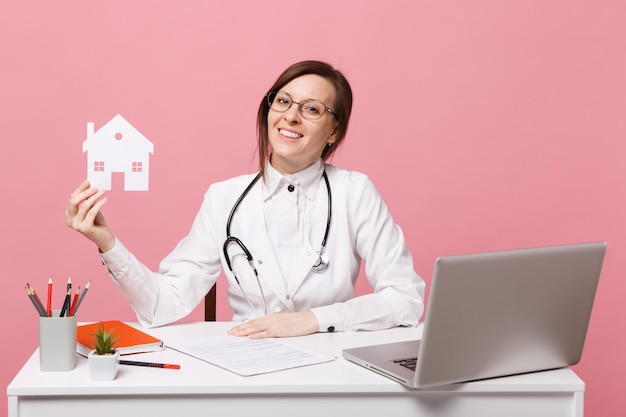 Médica sentar na mesa de trabalho no computador com documento médico segurar a casa no hospital isolado no fundo da parede rosa pastel. Mulher de estetoscópio de óculos de vestido médico. Conceito de medicina de saúde.