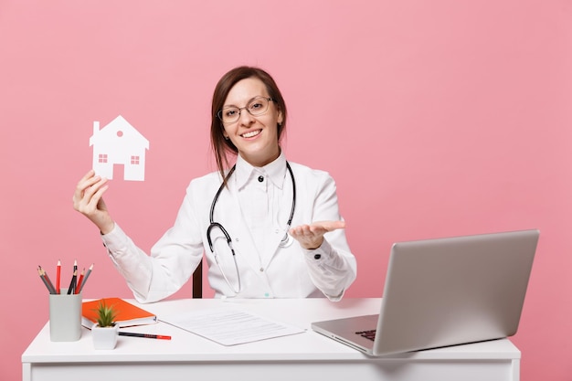 Médica sentar na mesa de trabalho no computador com documento médico segurar a casa no hospital isolado no fundo da parede rosa pastel. Mulher de estetoscópio de óculos de vestido médico. Conceito de medicina de saúde.