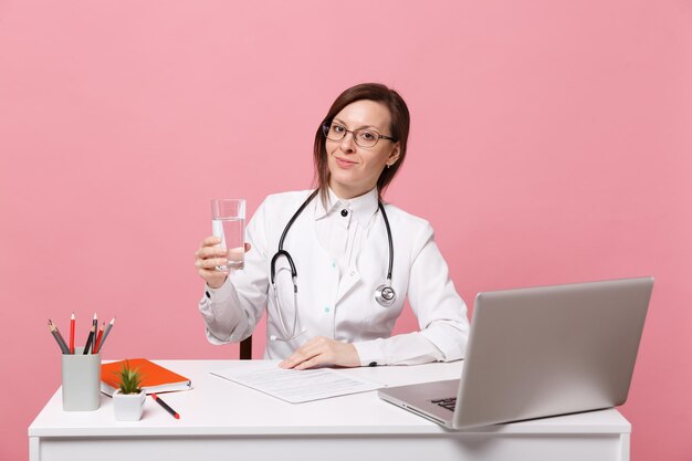 Médica sentar na mesa de trabalho no computador com documento médico segurar água no hospital isolado no fundo da parede rosa pastel. Mulher de estetoscópio de óculos de vestido médico. Conceito de medicina de saúde.