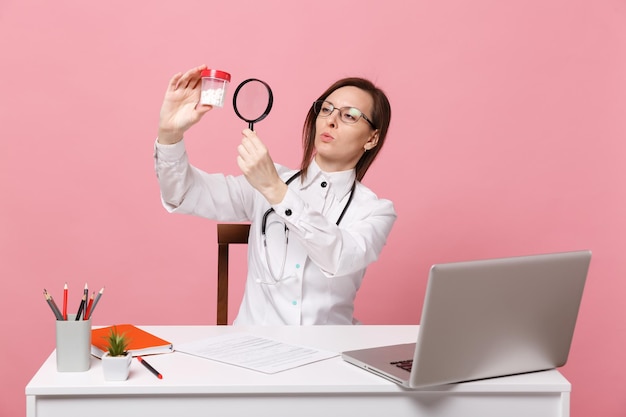 Médica sentar na mesa de trabalho no computador com documento médico segurar comprimidos no hospital, isolado no fundo da parede rosa pastel. mulher de estetoscópio de óculos de vestido médico. conceito de medicina de saúde.